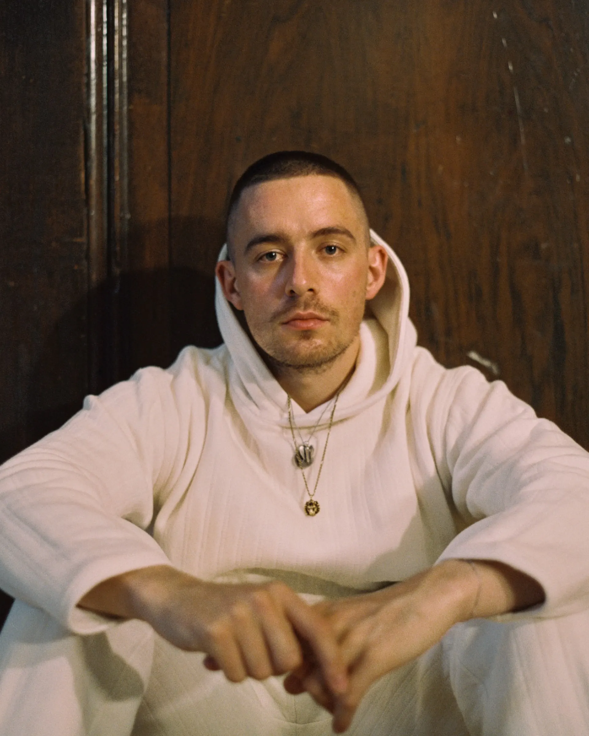 Dermot Kennedy sitting against a wooden door in a cream hoodie and joggers, arms resting on his knees