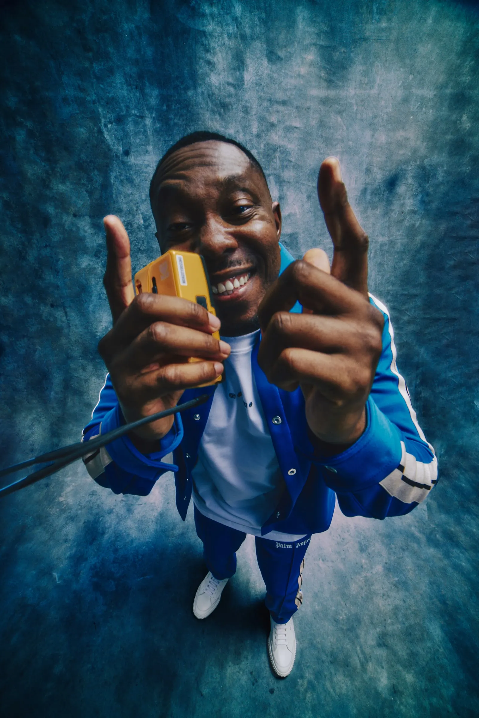 Dizzee Rascal grinning up at the camera in a blue track jacket, holding a yellow Walkman