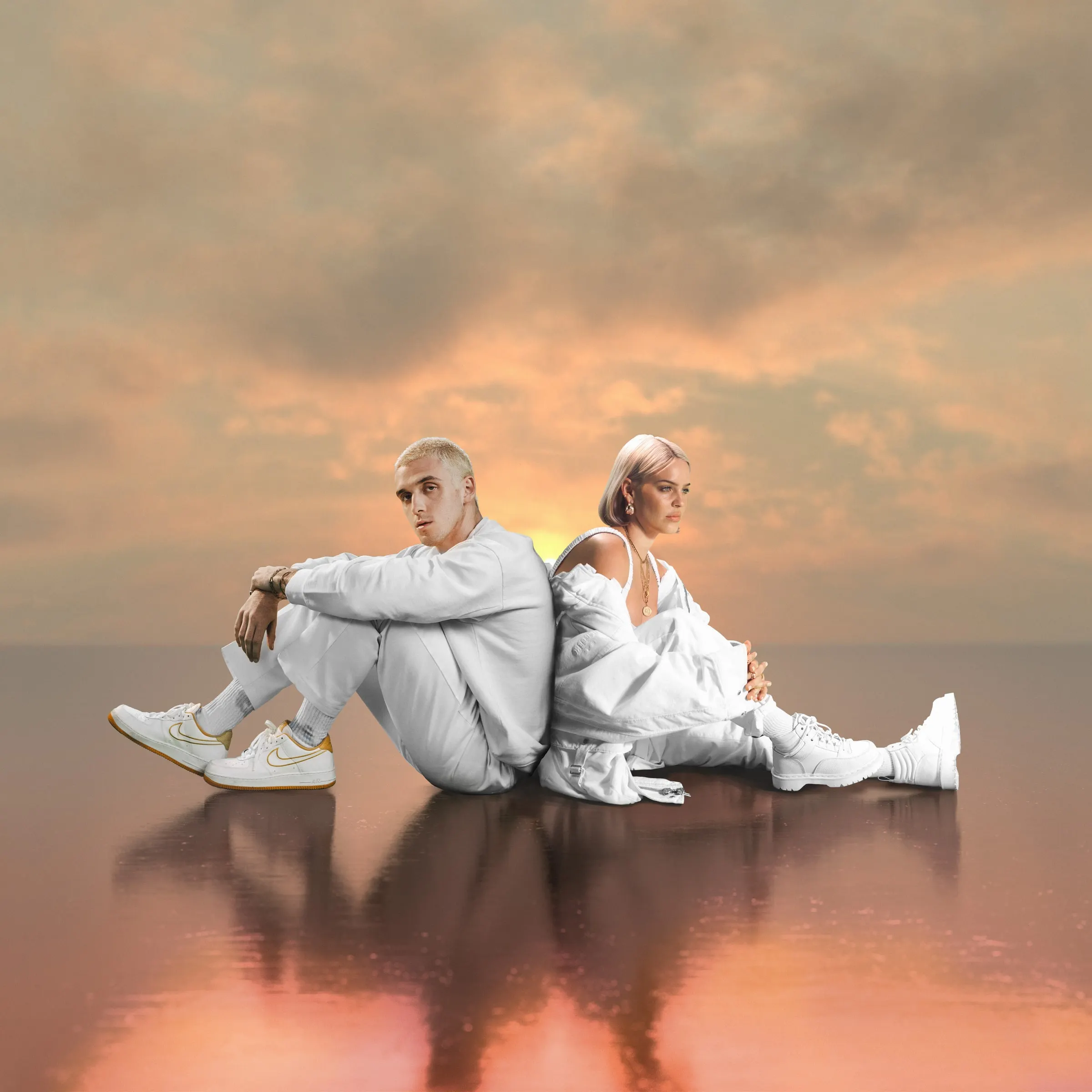 Lauv and Anne-Marie in white, sitting back to back on a reflective surface beneath a peach sunset sky