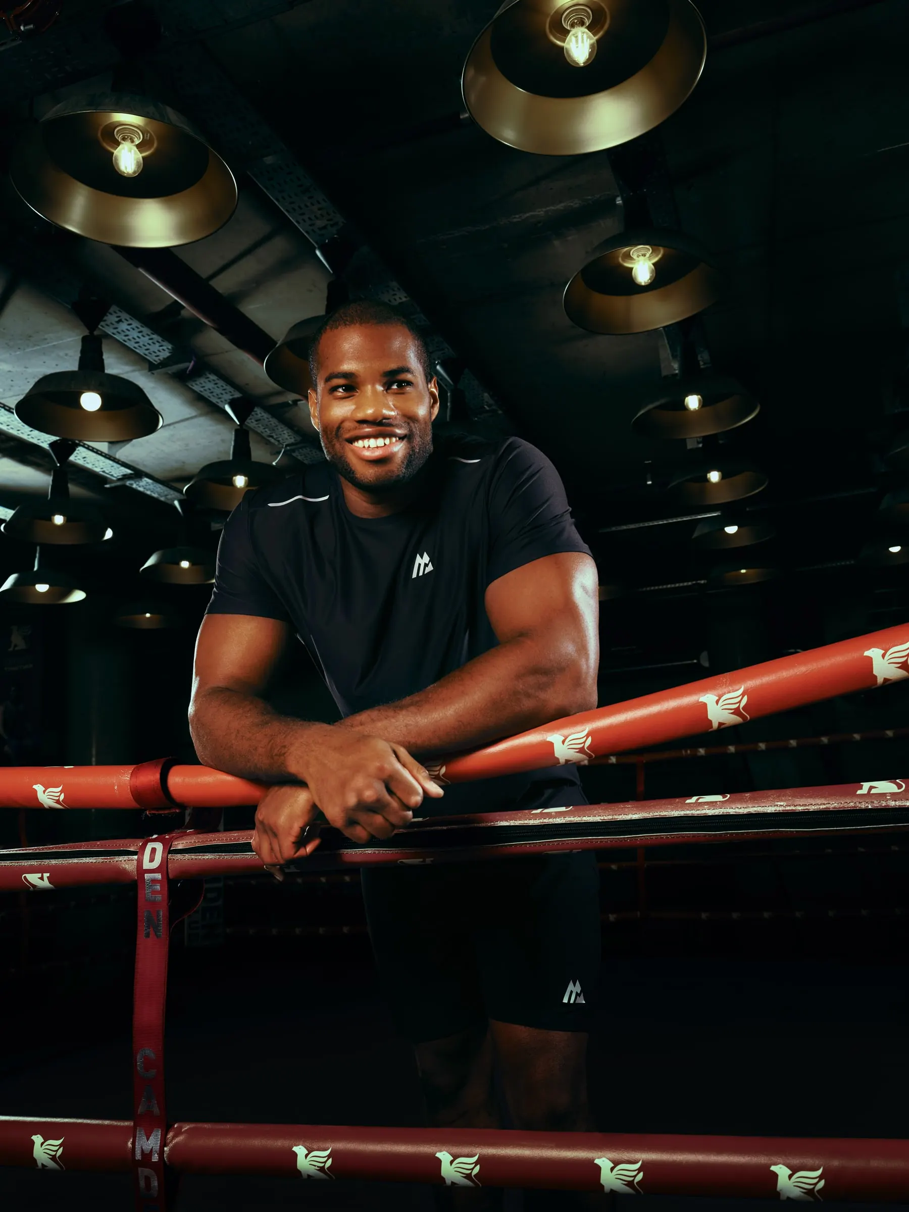 Daniel Dubois smiling, leaning on red ring ropes beneath industrial lamps