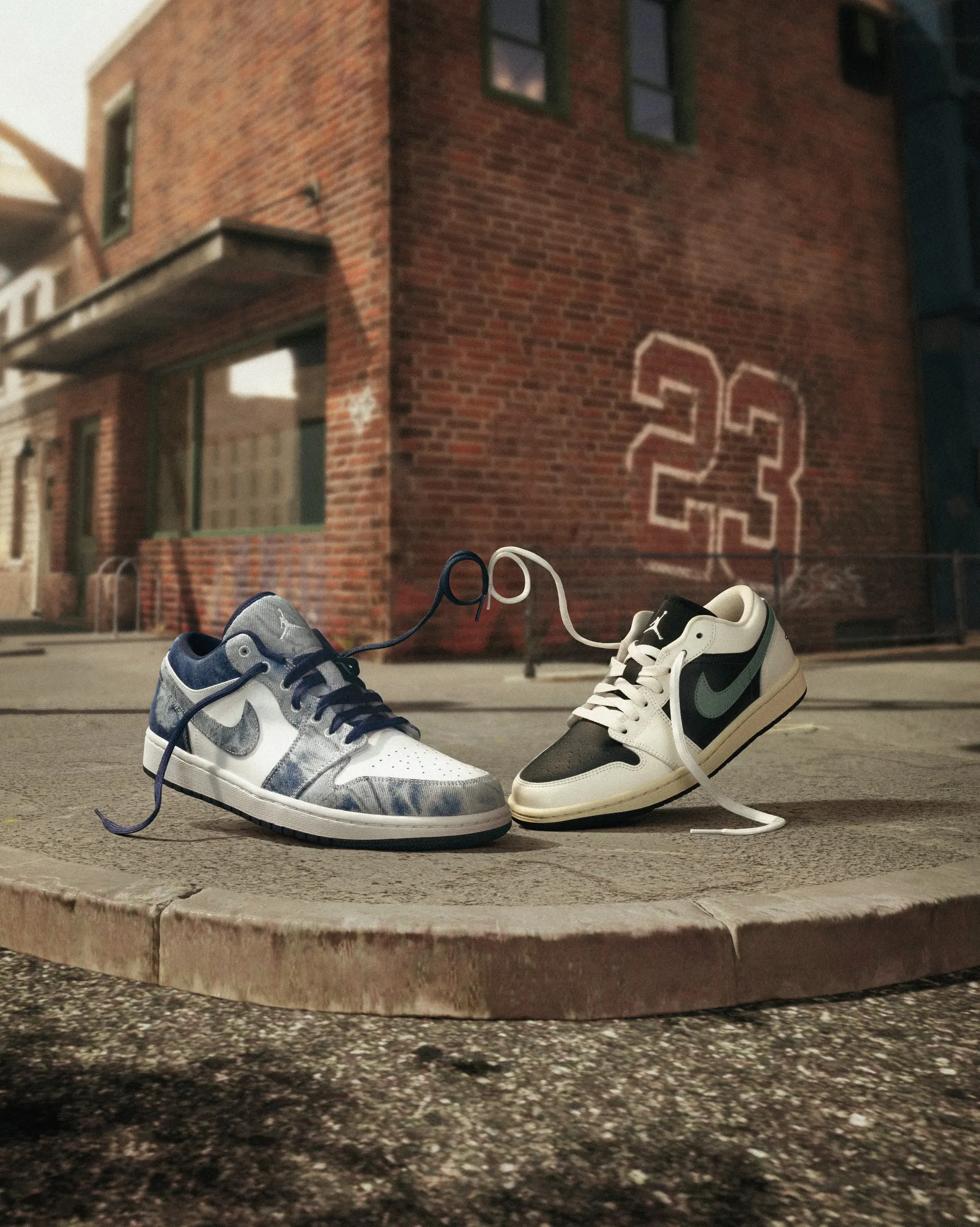 Two Air Jordan 1 Lows on a brick street corner, a 23 painted on the wall behind