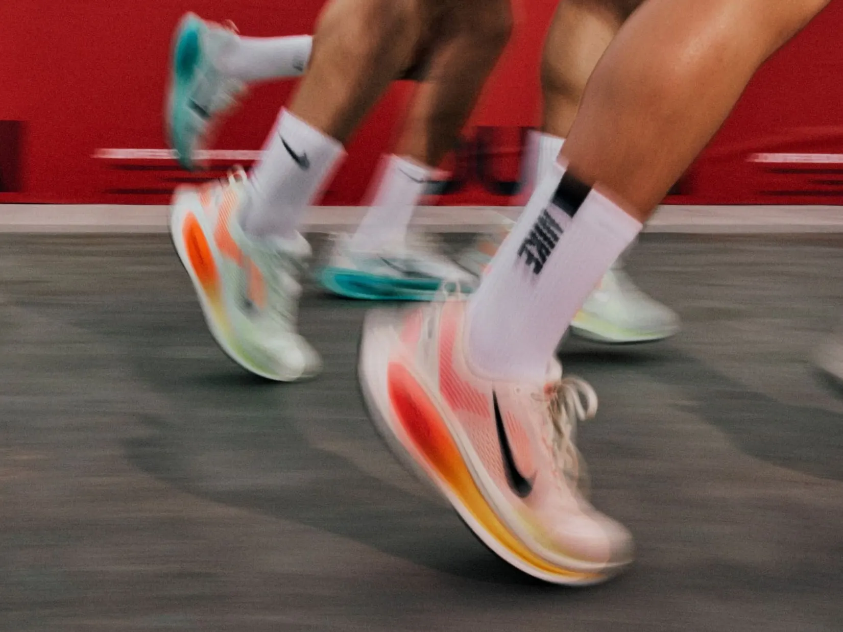 Runners in Vomero 18s blurred mid-stride against a red Nike wall