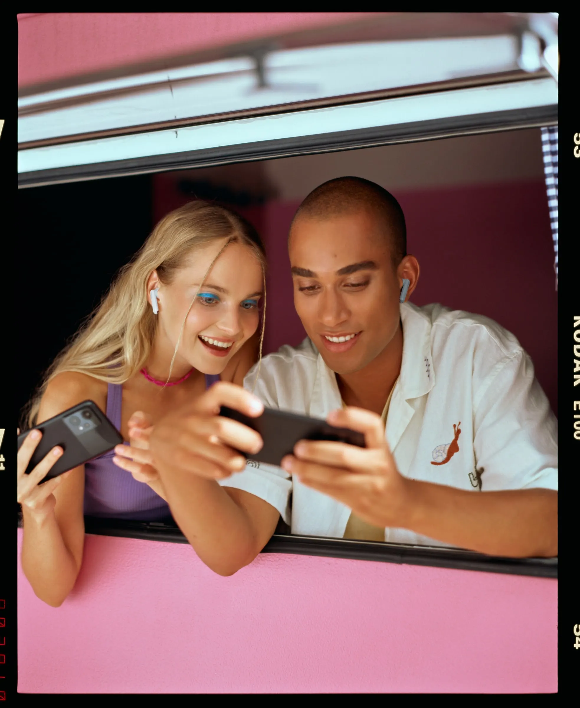 Two friends with earbuds leaning out of a pink van window, laughing at their phones