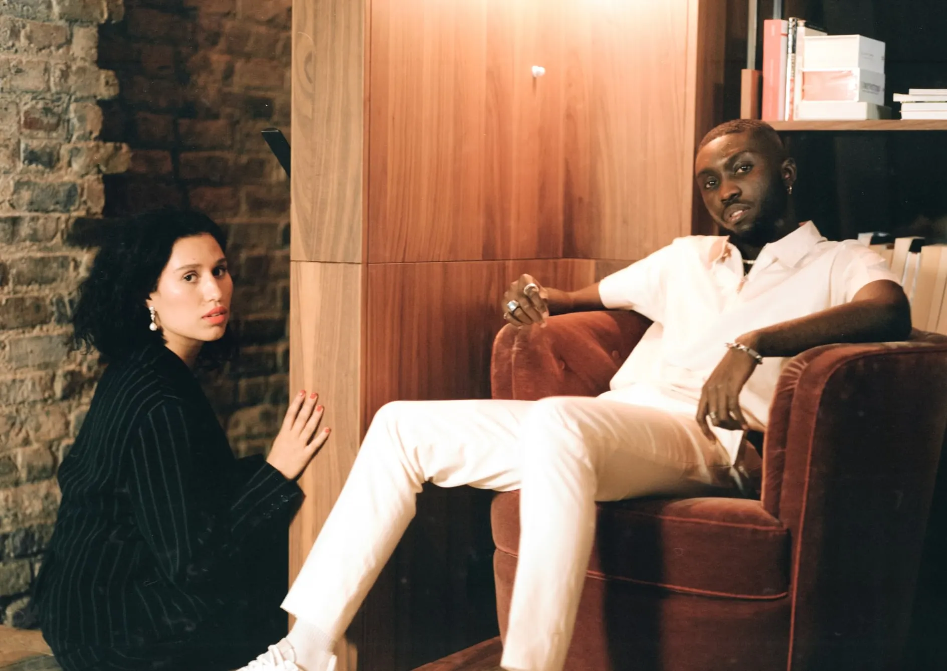 Odunsi in cream lounging in a velvet armchair, RAYE in black pinstripe sitting on the floorboards beside a brick wall